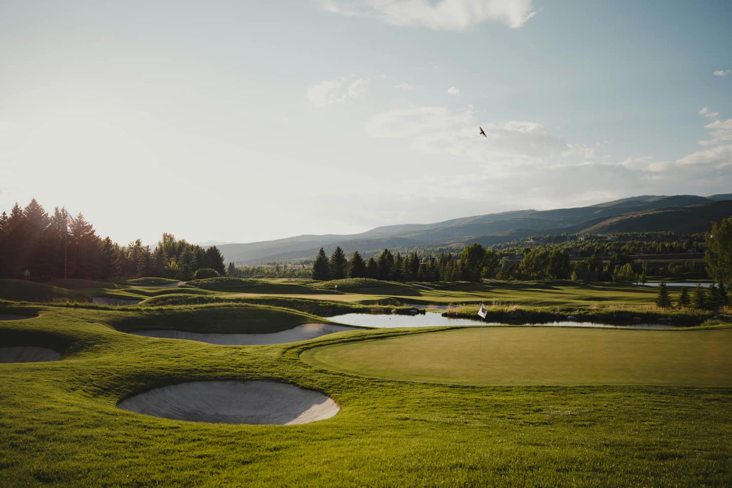 A golf green with bunkers and a flag, a pond and mountains behind
